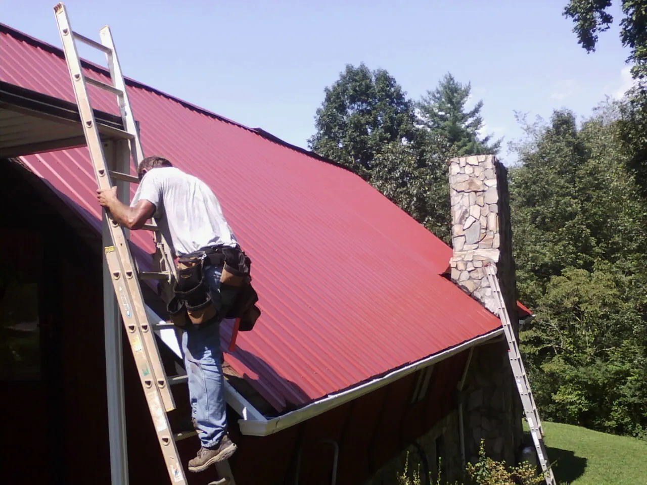 Roofer on a ladder beside a red metal roof and stone chimney — Soffit & Fascia Repair by Roofing Brothers in Goodhue